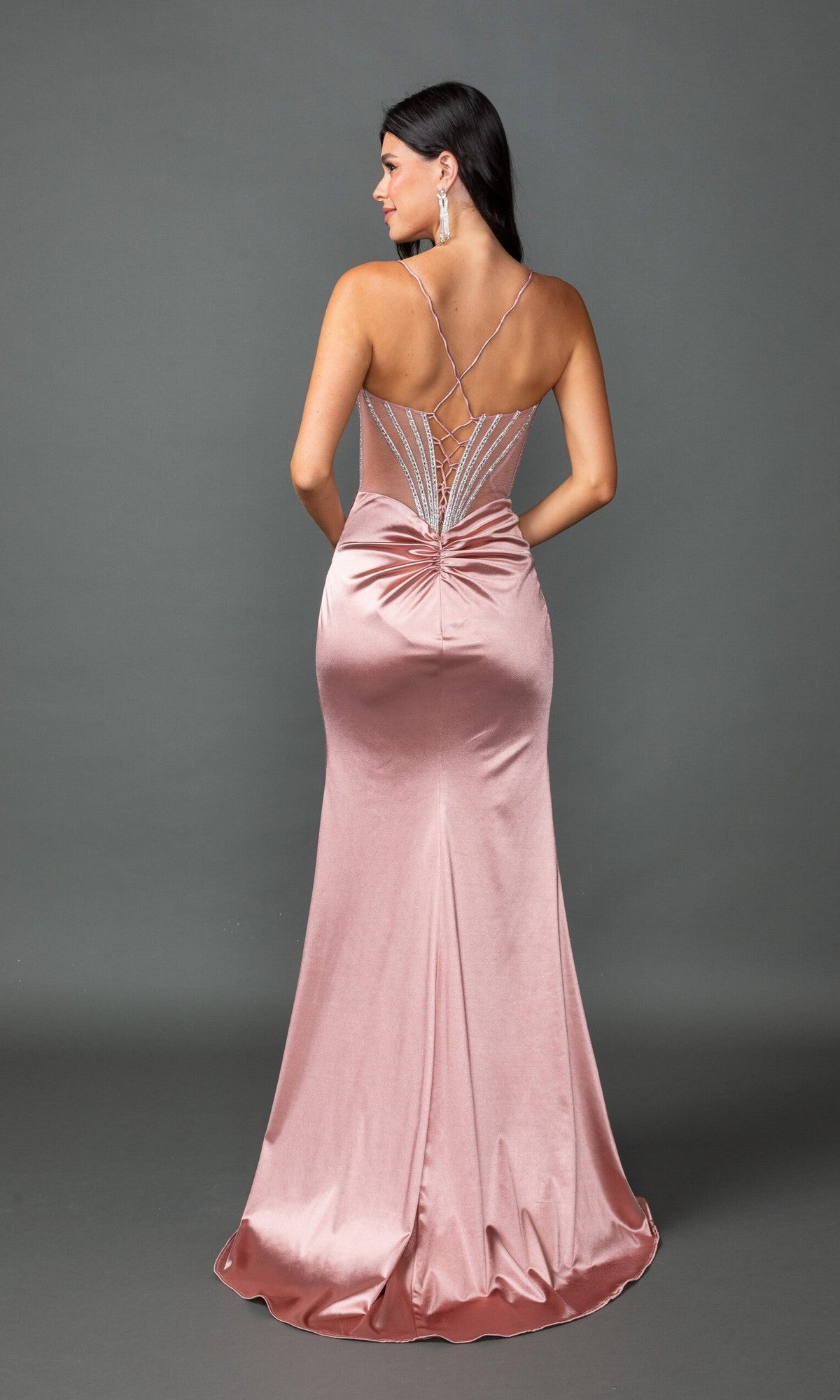 A woman wears the Long Prom Dress: Dancing Queen 4538, a pink satin beaded gown with a fitted silhouette, ruched lower back, and strappy lace-up back, standing against a gray background.